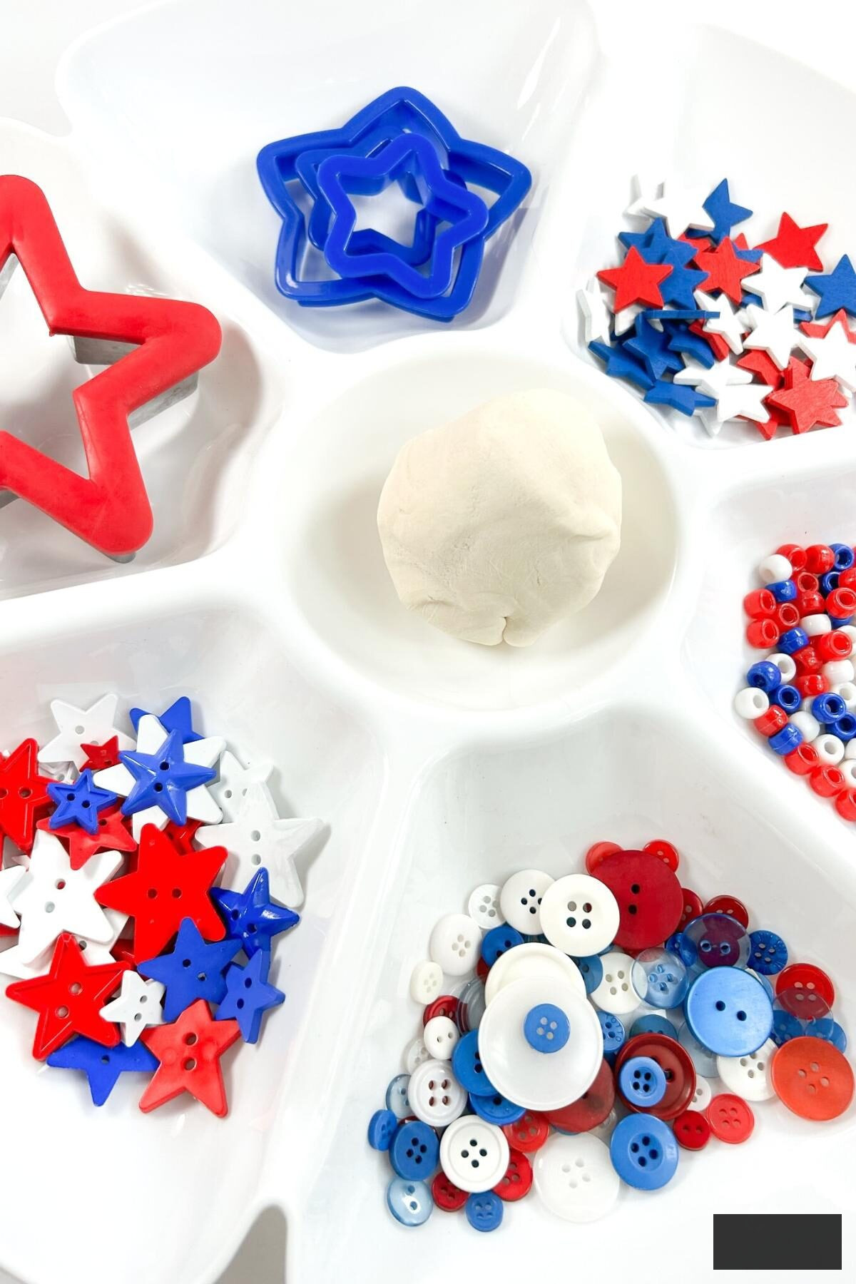 Patriotic playdough invitation arranged on a dip tray, ready for preschoolers to engage. Includes white homemade playdough, red, white, and blue stars, buttons, and cookie cutters.