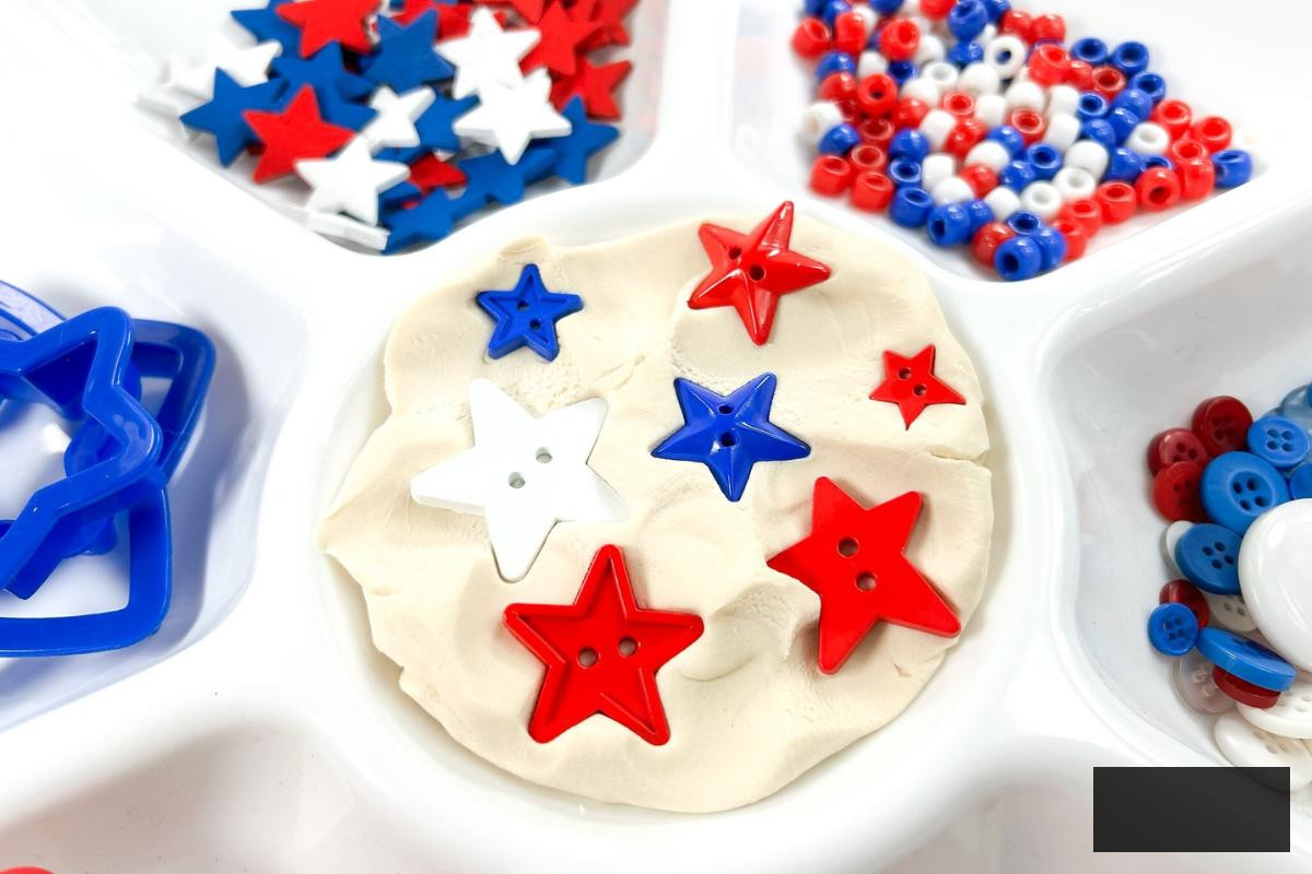 Red, white, and blue stars of varying sizes positioned in white playdough on a divided tray set up as a quick sensory activity for the 4th of July for children.
