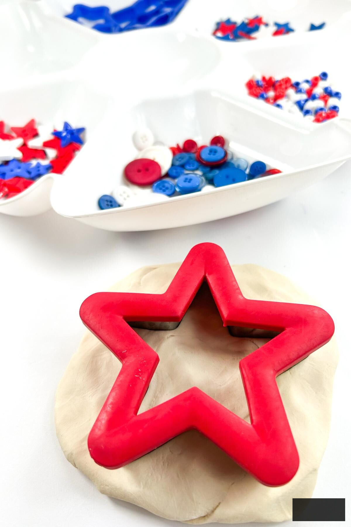 A red cookie cutter star cutting out white playdough with a tray filled with red, white, and blue buttons and stars in the background.