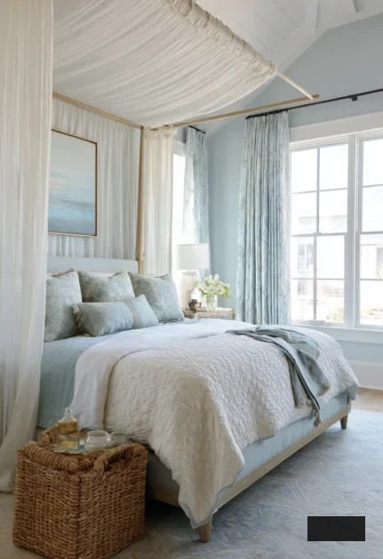 A tranquil coastal bedroom featuring a canopy bed draped in white fabric, light blue and white bedding, multiple cushions, a wicker basket at the foot, expansive windows adorned with patterned curtains, and a coastal artwork above the headboard.