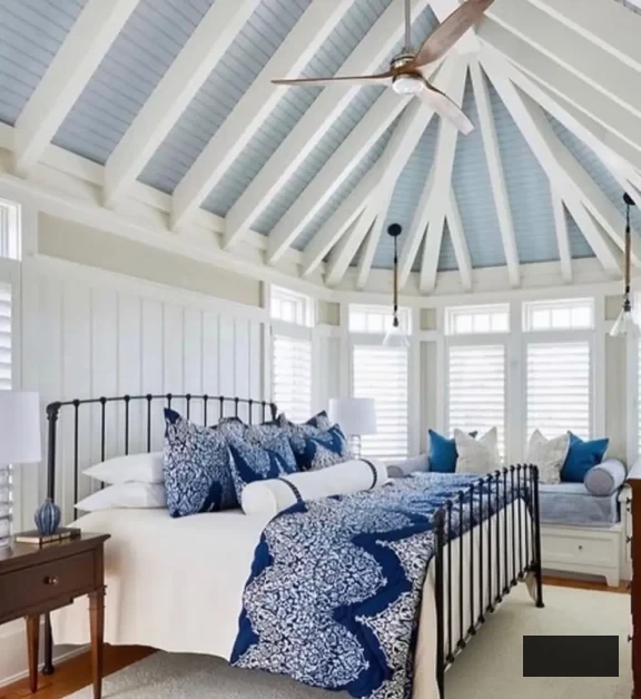A radiant coastal bedroom with a lofty, vaulted ceiling and exposed beams. The bed features blue and white bedding, accompanied by a window seat with cushions. Ample windows and white walls fill the room with natural light.