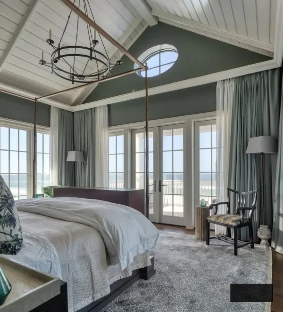 A bright, spacious coastal bedroom showcasing a vaulted ceiling, canopy bed, expansive windows, and doors leading to a patio, teal walls, light curtains, a grey rug, and a blend of modern and coastal decor.
