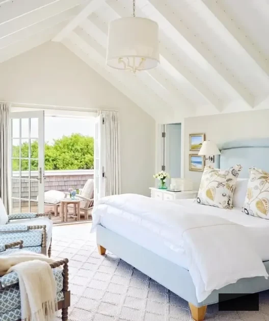 Bright, airy coastal bedroom showcasing a white vaulted ceiling, light blue bed, floral cushions, two armchairs, and glass doors leading to a sunlit balcony with outdoor seating and lush greenery beyond.