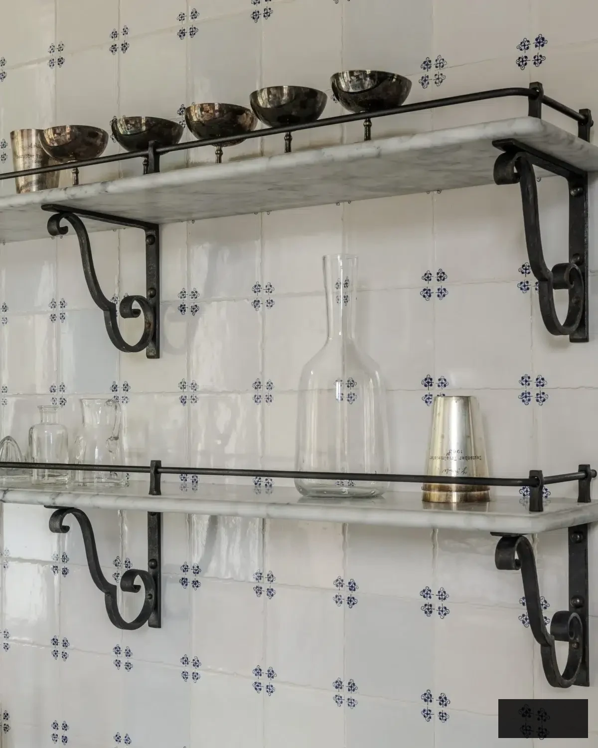 Zoomed-in view of wrought-iron supports holding up Carrara marble shelves against hand-painted white tiles adorned with tiny blue designs. The shelves present silver bowls, glass carafes, and vintage barware — a flawless blend of rustic Italian craftsmanship and refined sophistication.