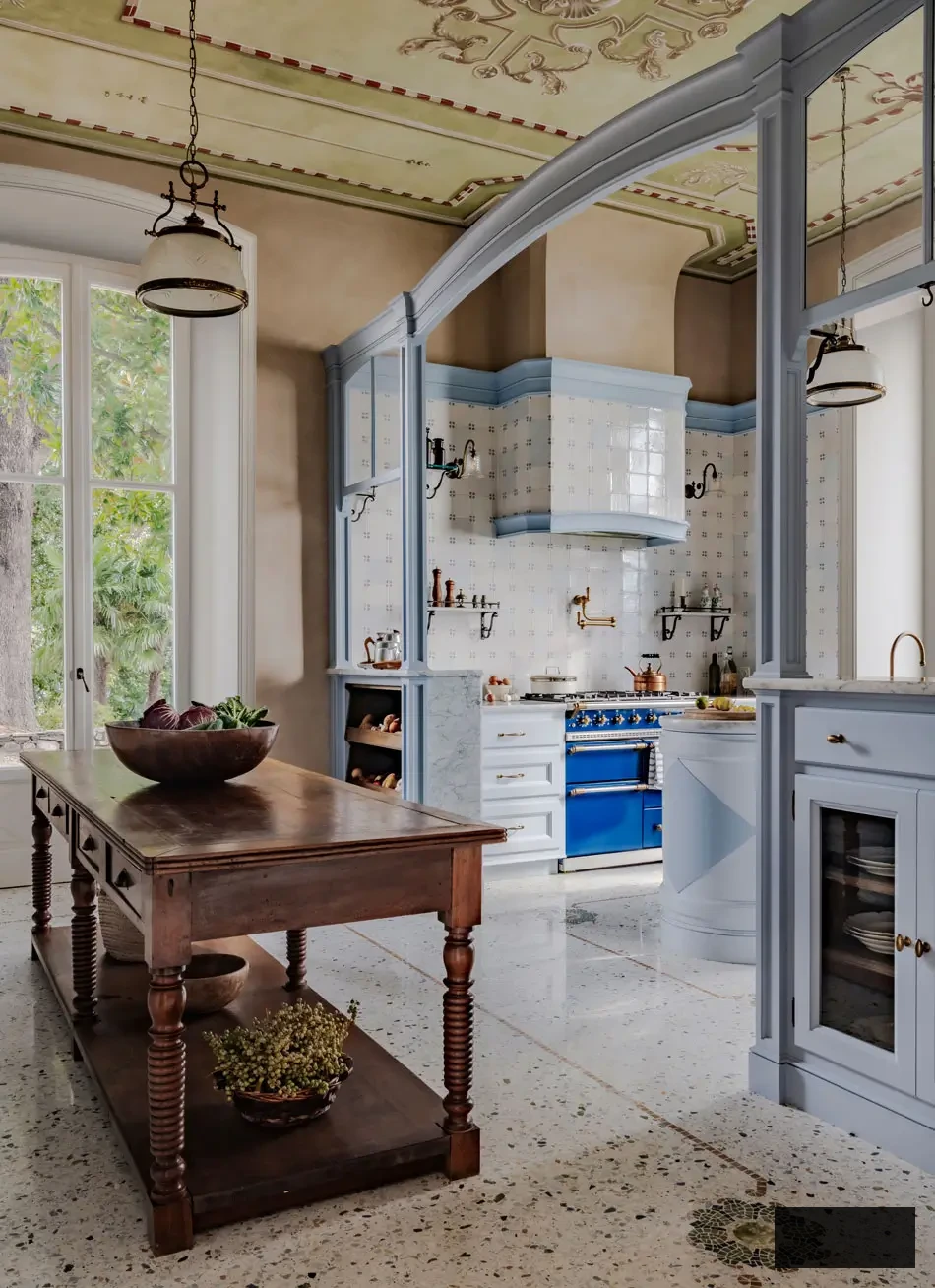 Open-layout view of Artichoke’s custom Lake Maggiore kitchen, showcasing an arched joinery frame, sky-blue cabinetry, marble island, and vintage wooden prep table. The area merges classic Italian detailing with a soft, layered color scheme for an everlasting lakeside ambiance.
