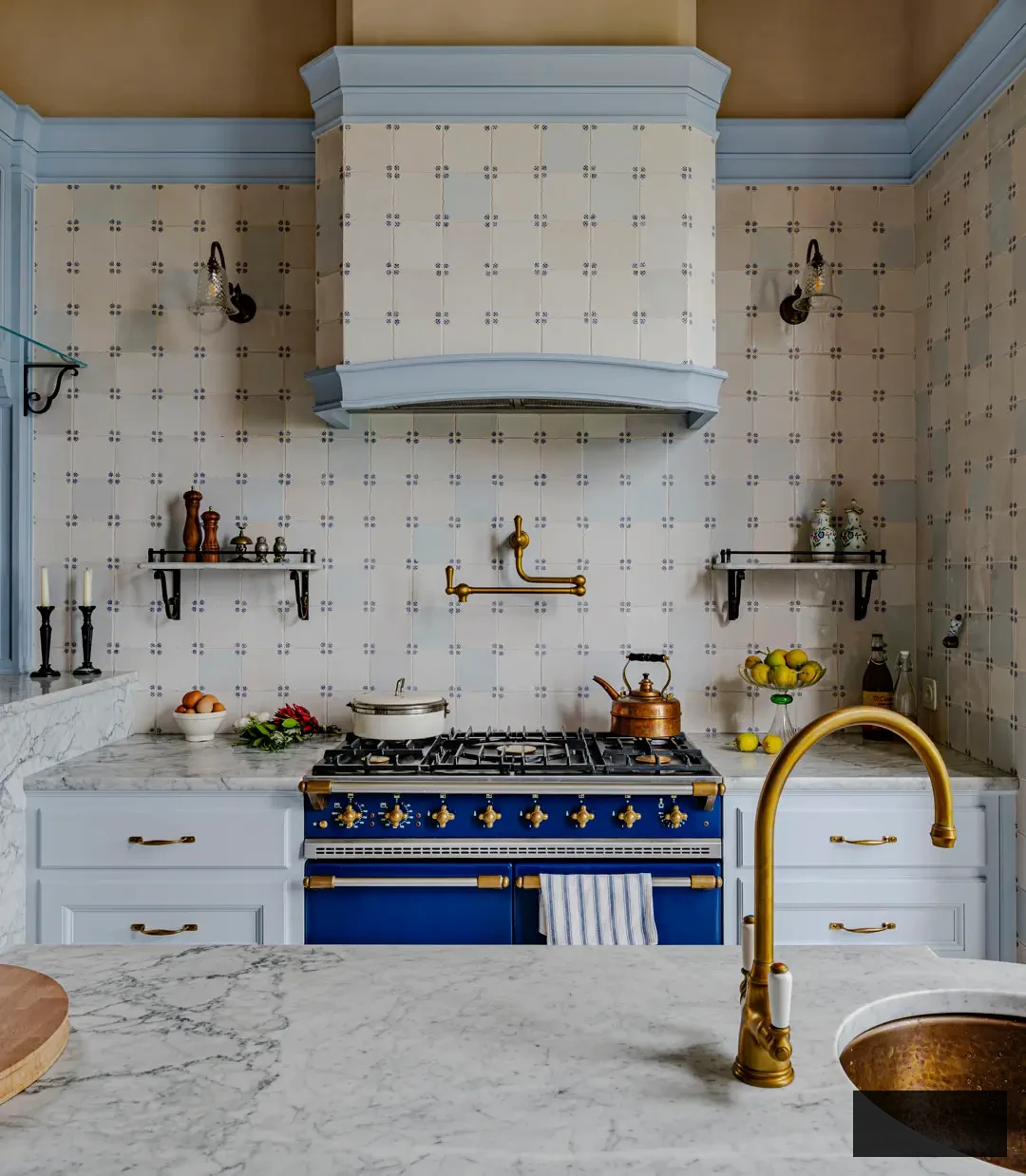 Marble countertops adorned with fluted edge detail, brass faucet, and deep farmhouse sink in a handcrafted Italian kitchen designed by Artichoke. The arrangement showcases classic tiled walls, polished brass fixtures, and the sophistication of soft-blue painted cabinetry against neutral plaster walls.