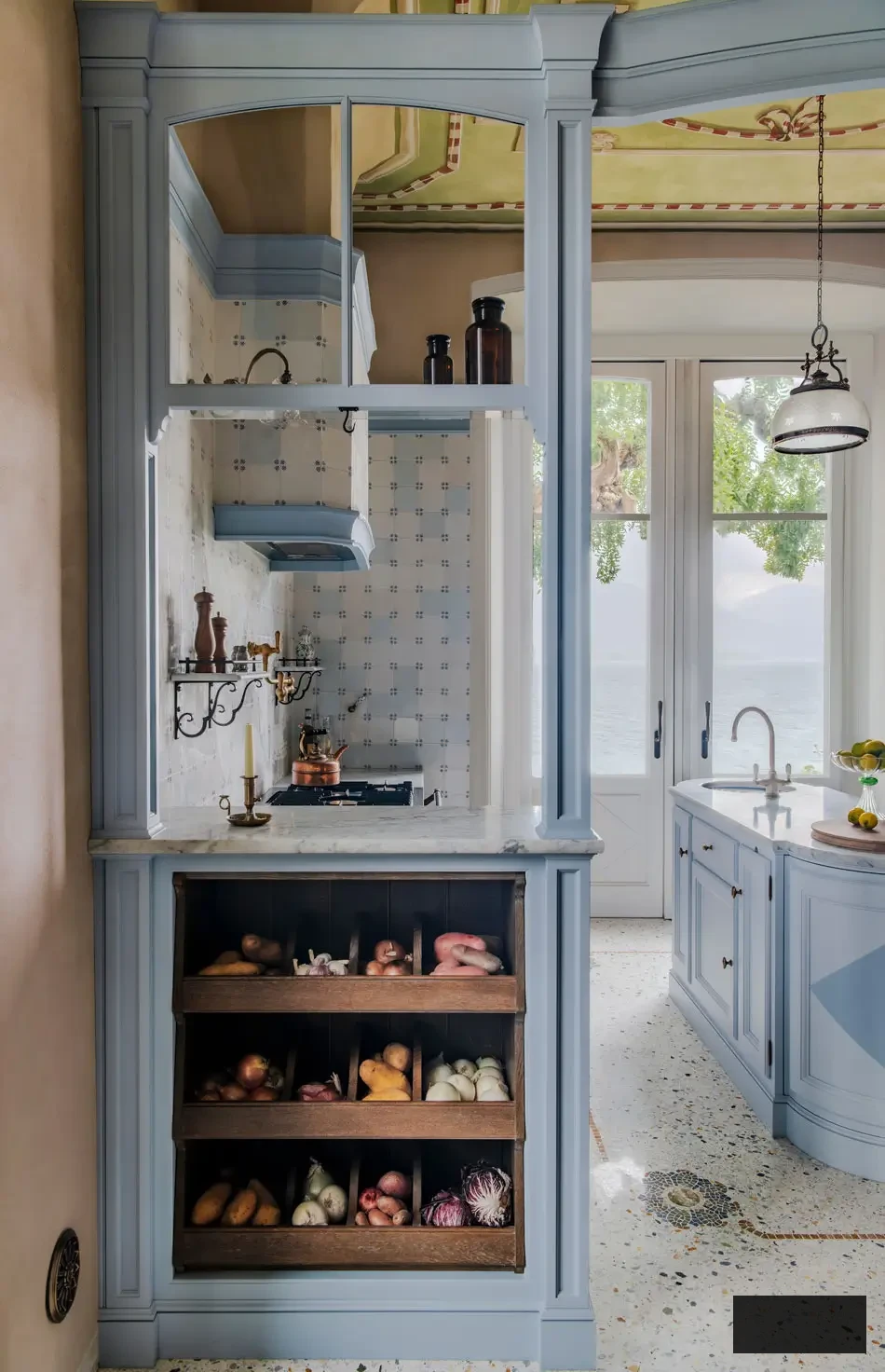 Ingenious integrated vegetable drawers crafted from oak and framed by blue-grey cabinetry in an Italian villa kitchen. Through the marble-trimmed partition, the view reveals the island sink and panoramic Lake Maggiore vista beyond, merging function and beauty flawlessly.