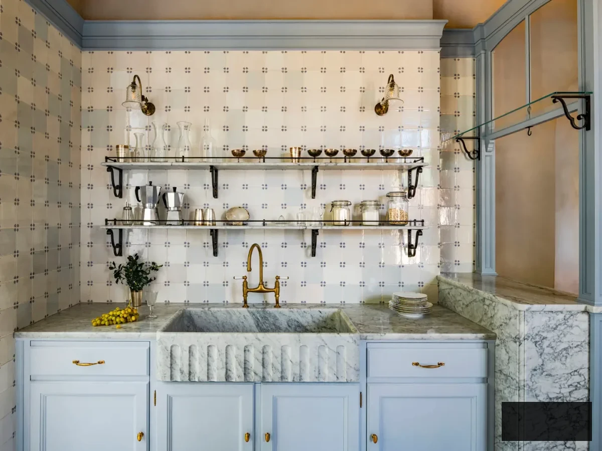 Front view of fluted marble farmhouse sink with brass bridge faucet, open glass shelving, and handcrafted ceramic tiles in an Artichoke custom kitchen on Lake Maggiore. Glass carafes, brass bowls, and gentle lighting enhance the artisanal, lived-in luxurious ambiance of the space.