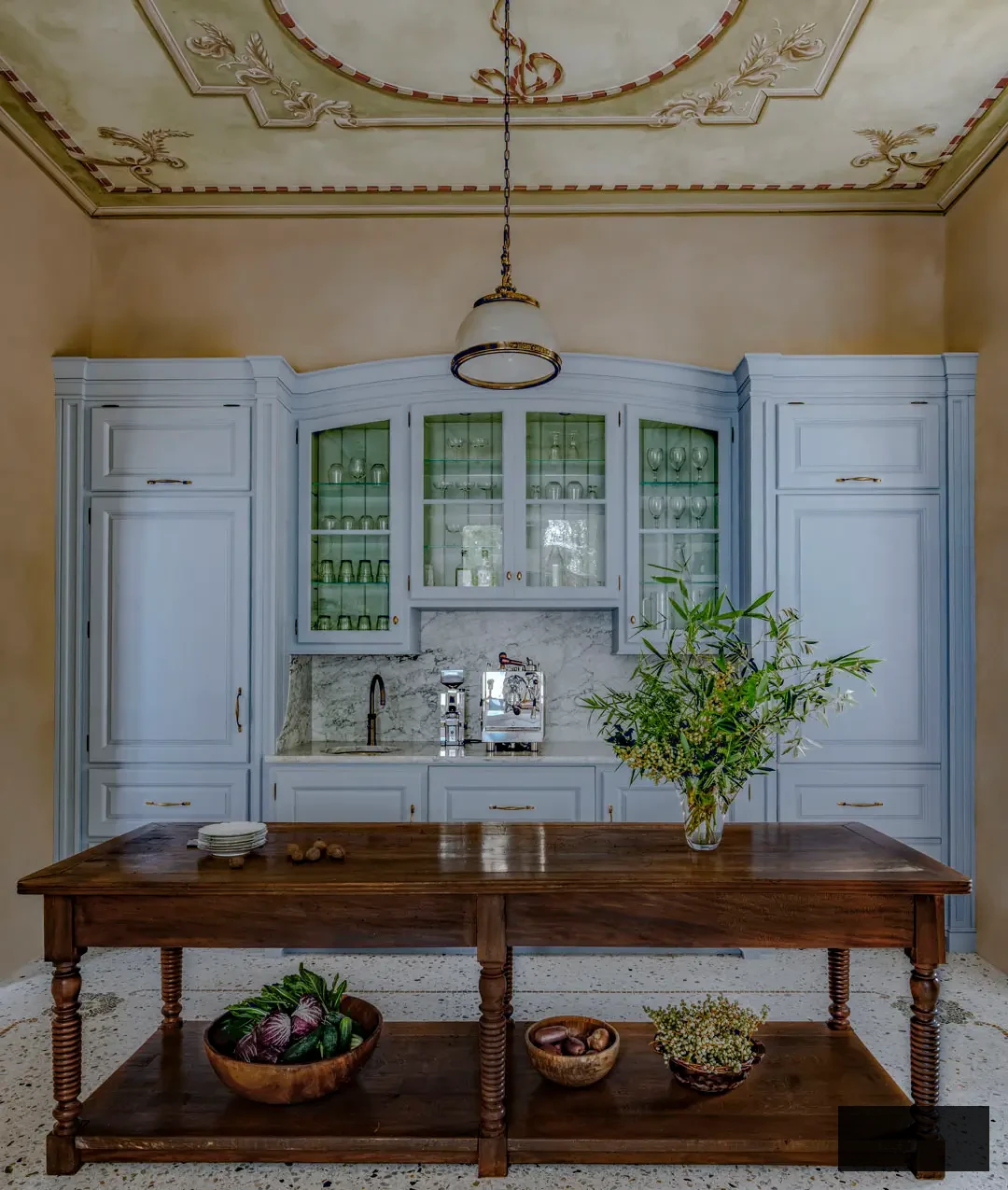Back wall of the Lake Maggiore villa kitchen displaying symmetrical cabinetry with arched glass doors, marble backsplash, and a built-in coffee nook. The antique wooden island introduces warmth and contrast to the powder-blue cabinetry and the intricate hand-painted ceiling above.