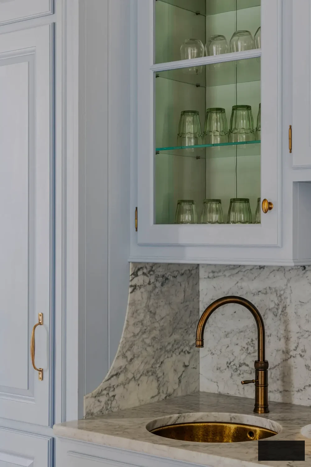 Intricate view of marble countertop with rounded splashback and vintage brass sink in a custom Italian kitchen by Artichoke. The subdued blue cabinetry, glass shelving, and antiqued brass hardware reflect the villa’s historic allure while upholding contemporary craftsmanship precision.