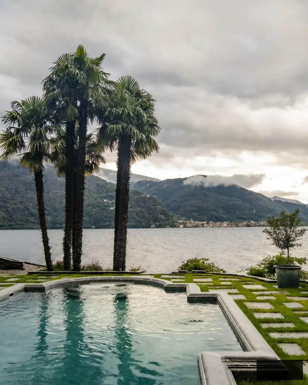 Picturesque view of Lake Maggiore framed by palm trees adjacent to a stone-edged swimming pool, with mist-covered peaks in the distance. The vista captures the laid-back elegance of the Italian Riviera, where traditional garden design intersects with natural lakeside beauty beneath a soft, cloud-laden sky.