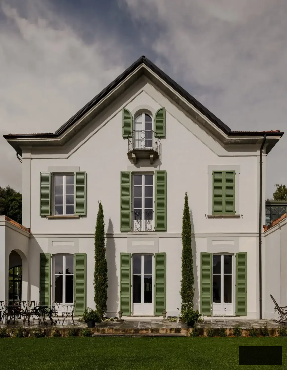 Sophisticated white Italian villa on the banks of Lake Maggiore featuring green shutters, tall cypress trees, and a stone terrace. The 19th-century façade reflects neoclassical elegance, surrounded by manicured gardens and wrought-iron outdoor furniture — a tranquil lakeside getaway that embodies timeless Italian architecture and subtle luxury.