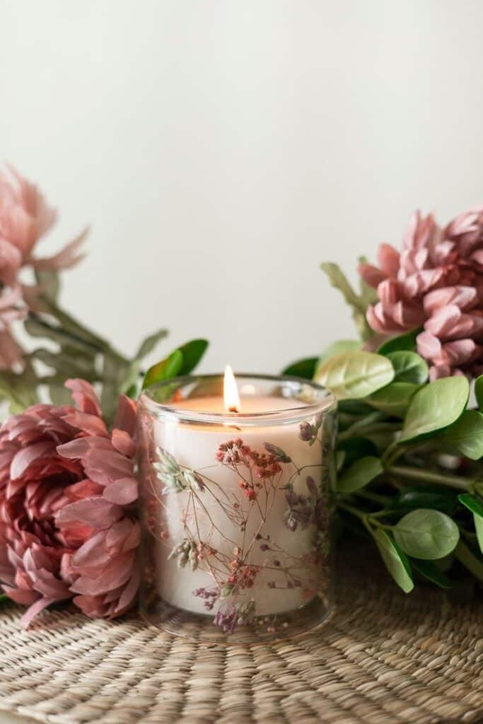 Illuminated botanical candle accompanied by dried flowers and green leaves on a wicker surface
