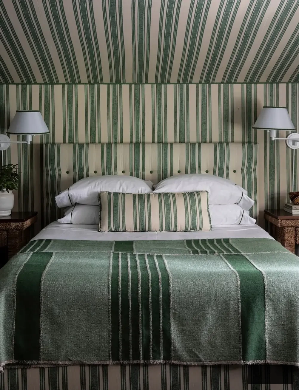 Cozy guest bedroom by Mark D. Sikes featuring green-and-white striped wallpaper, a matching upholstered headboard, and layered striped textiles.