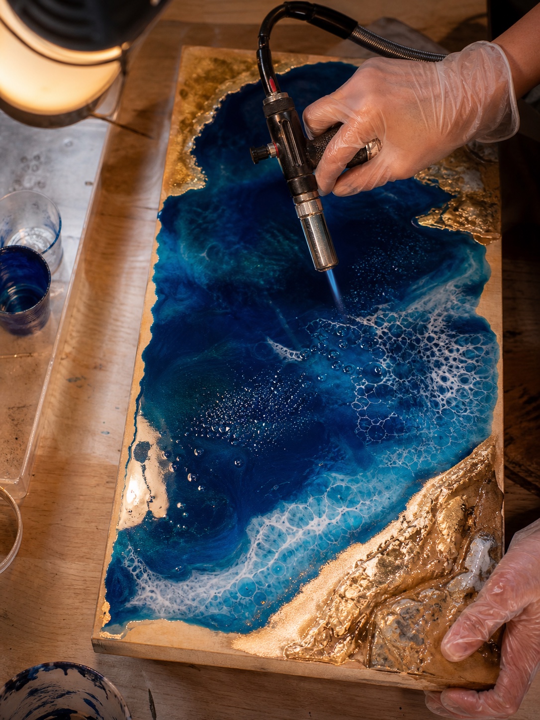 Resin art ocean pour bubble removal Gloved hands utilize a torch to eliminate bubbles from a blue and gold resin ocean pour. | Sky Rye Design Gloved hands utilize a torch to eliminate bubbles from a blue and gold resin ocean pour.