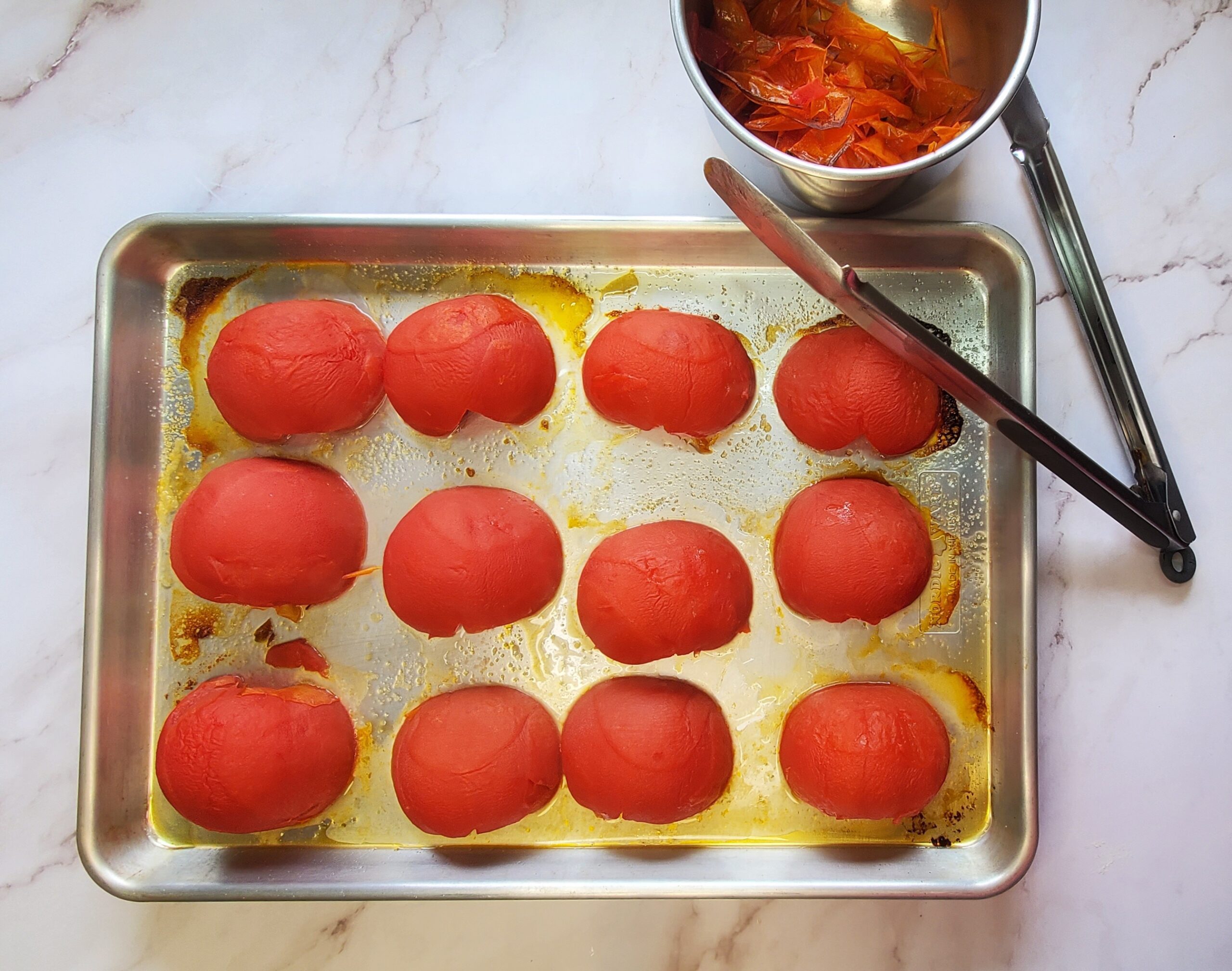 Tomates beefsteak assados com uma pinça descansando na assadeira, ao lado de uma tigela com peles de tomate.