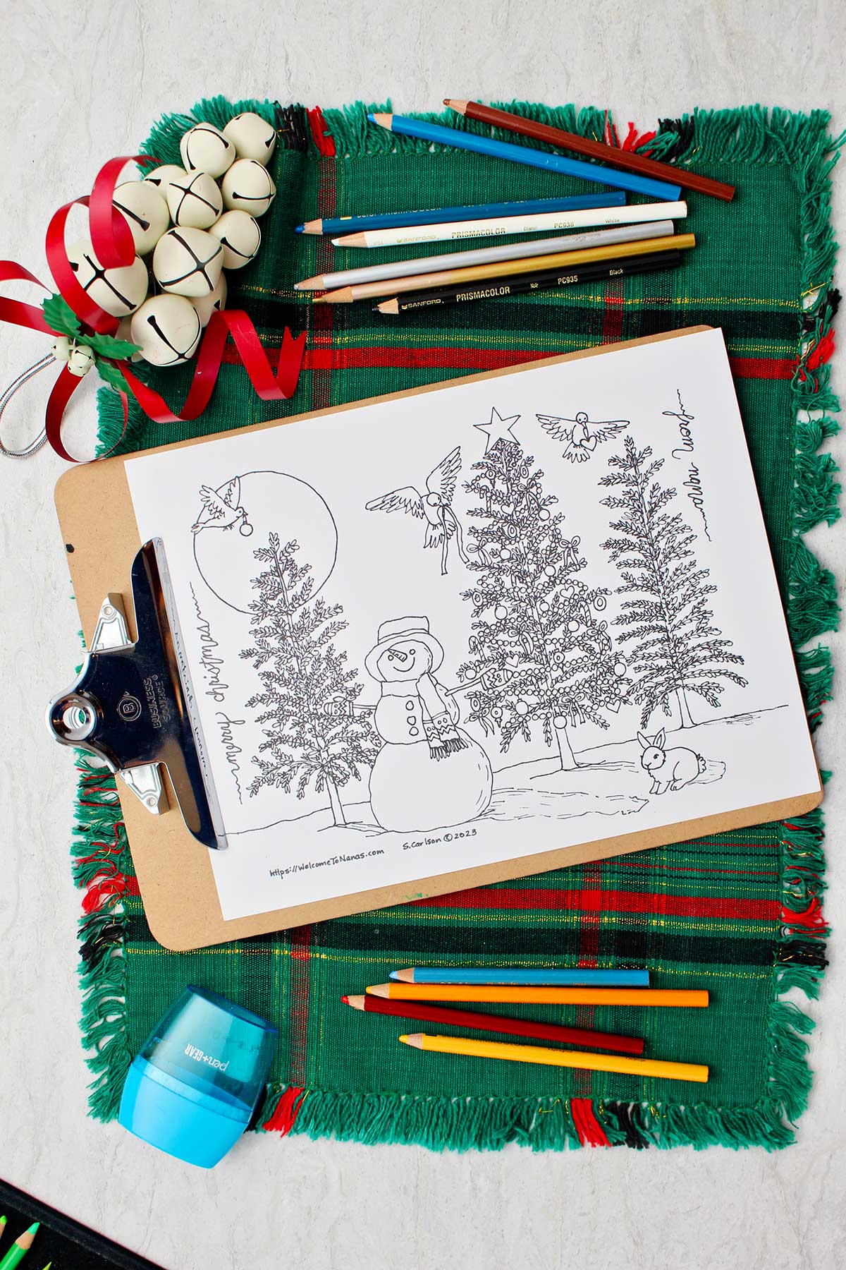Página de colorir árvore de natal sem cor em um clipboard descansando sobre um lugar de mesa verde e vermelho com lápis de cor, um apontador e sinos ao lado.
