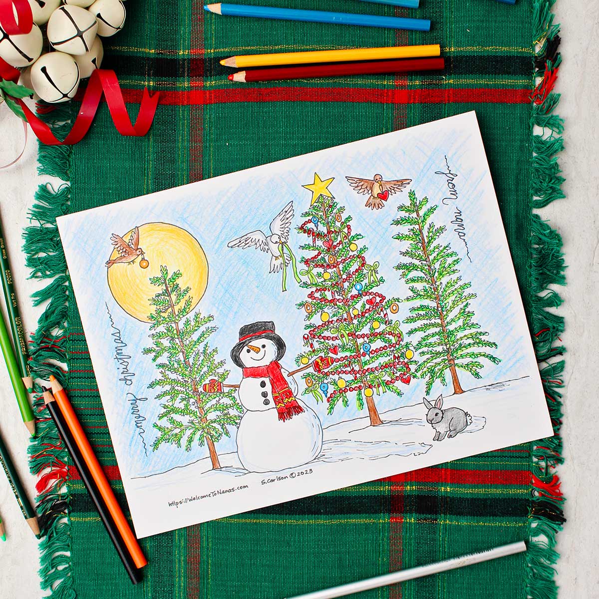Página de colorir de árvore de Natal completada descansando sobre um caminho de mesa verde e vermelho com sinos decorativos brancos por perto. Imagem quadrada.