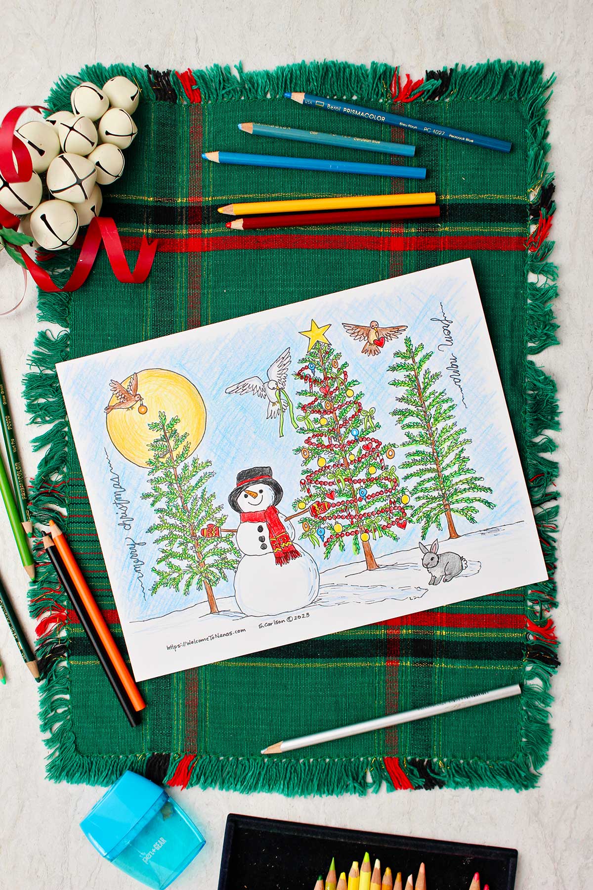 Página de colorir árvore de natal completa descansando sobre um lugar de mesa verde e vermelho com sinos decorativos brancos e lápis de cor ao lado.