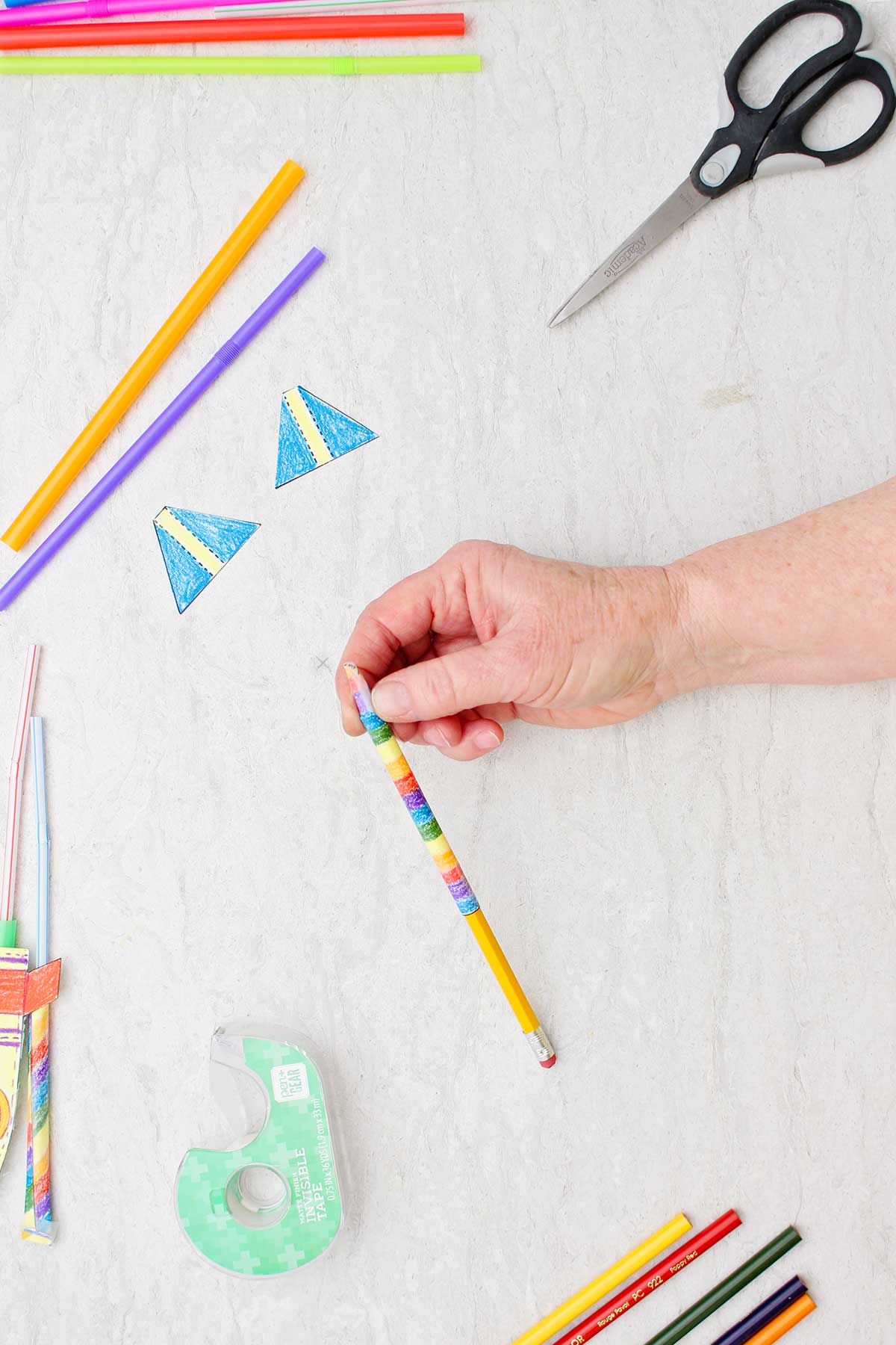Pessoa enrolando o retângulo colorido de papel em torno de um lápis com outros suprimentos ao redor.
