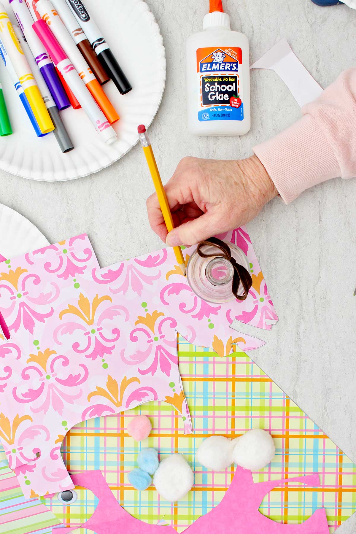 Pessoa usando uma garrafa de vidro para traçar um círculo em um pedaço de papel rosa estampado, com outros materiais próximos.