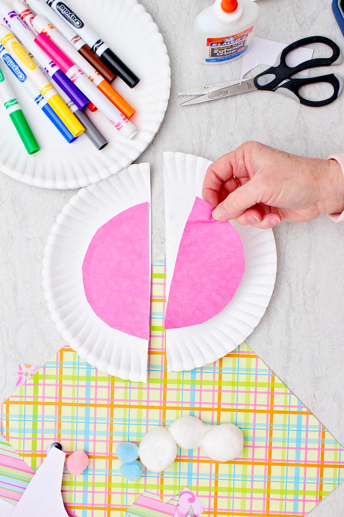 Pessoa colocando um corte em forma de meio círculo de papel rosa em uma metade de um prato grande para fazer orelhas de coelho, com outros materiais próximos.