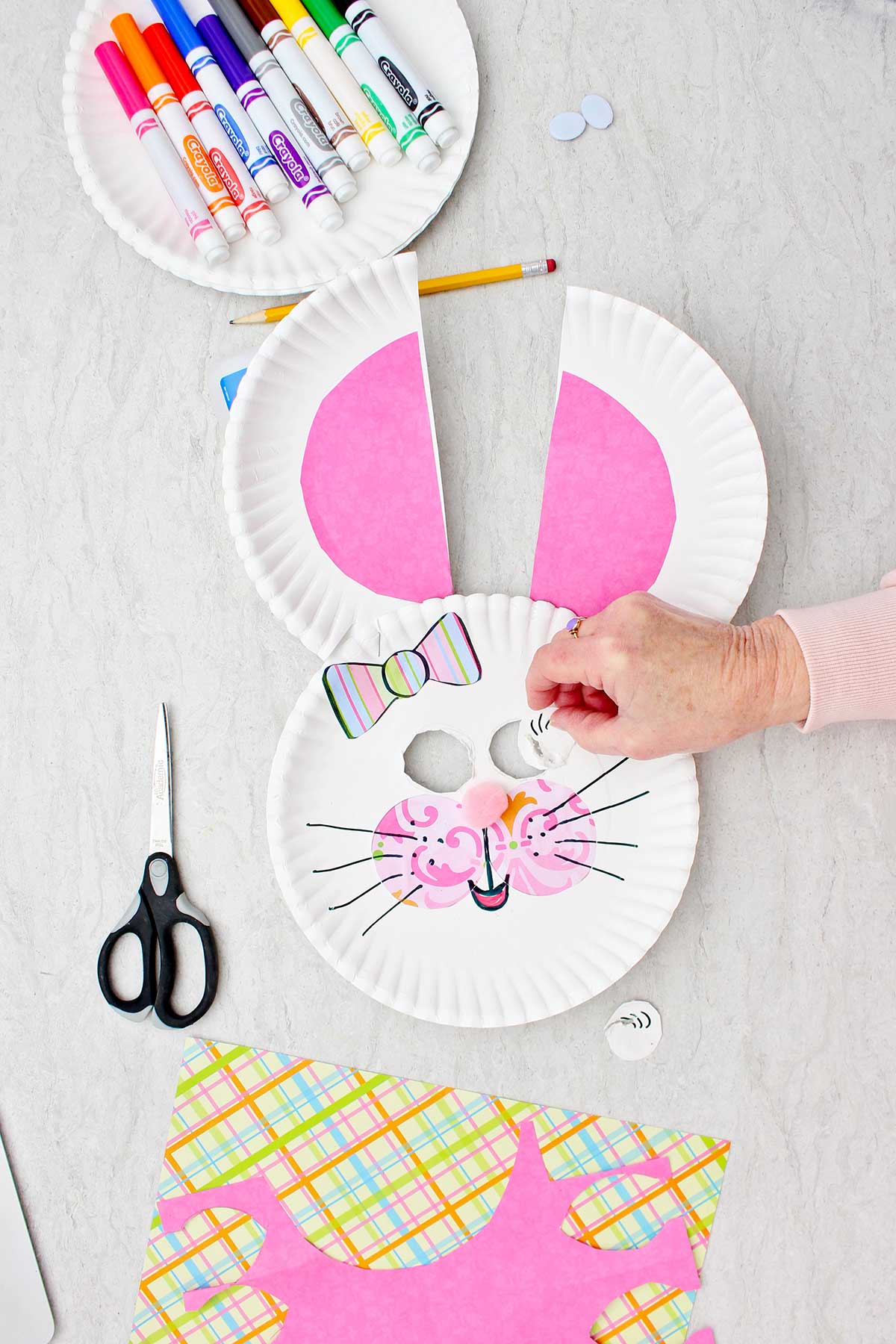 Pessoa recortando e removendo buracos para os olhos de um coelho de prato de papel para uma máscara com uma placa de marcadores e outros suprimentos nas proximidades.