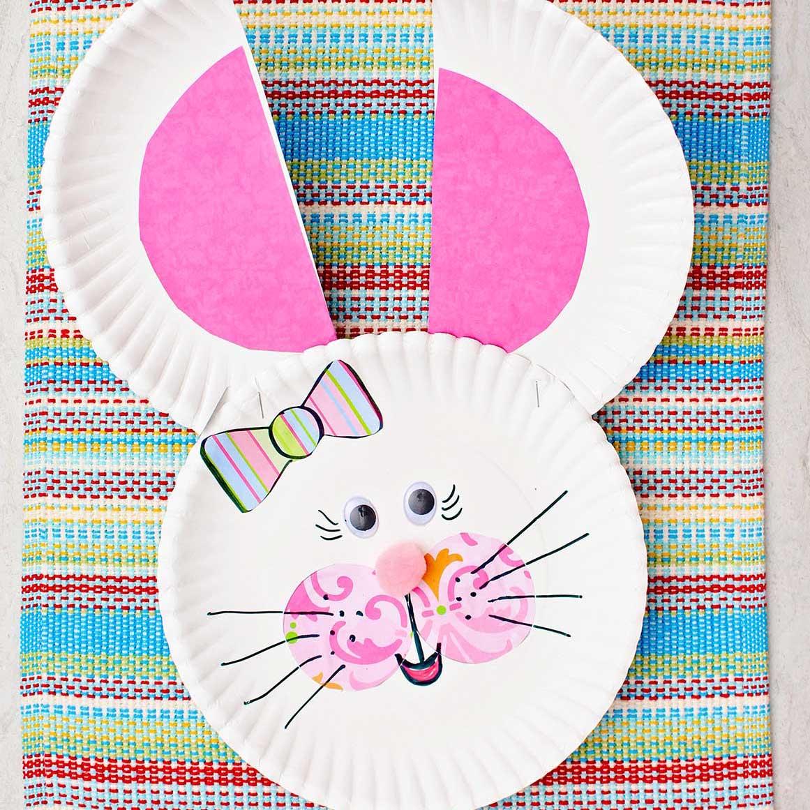 Coelhinho de prato de papel com orelhas e bochechas rosas, nariz de pom pom, laço listrado e olhos googly.