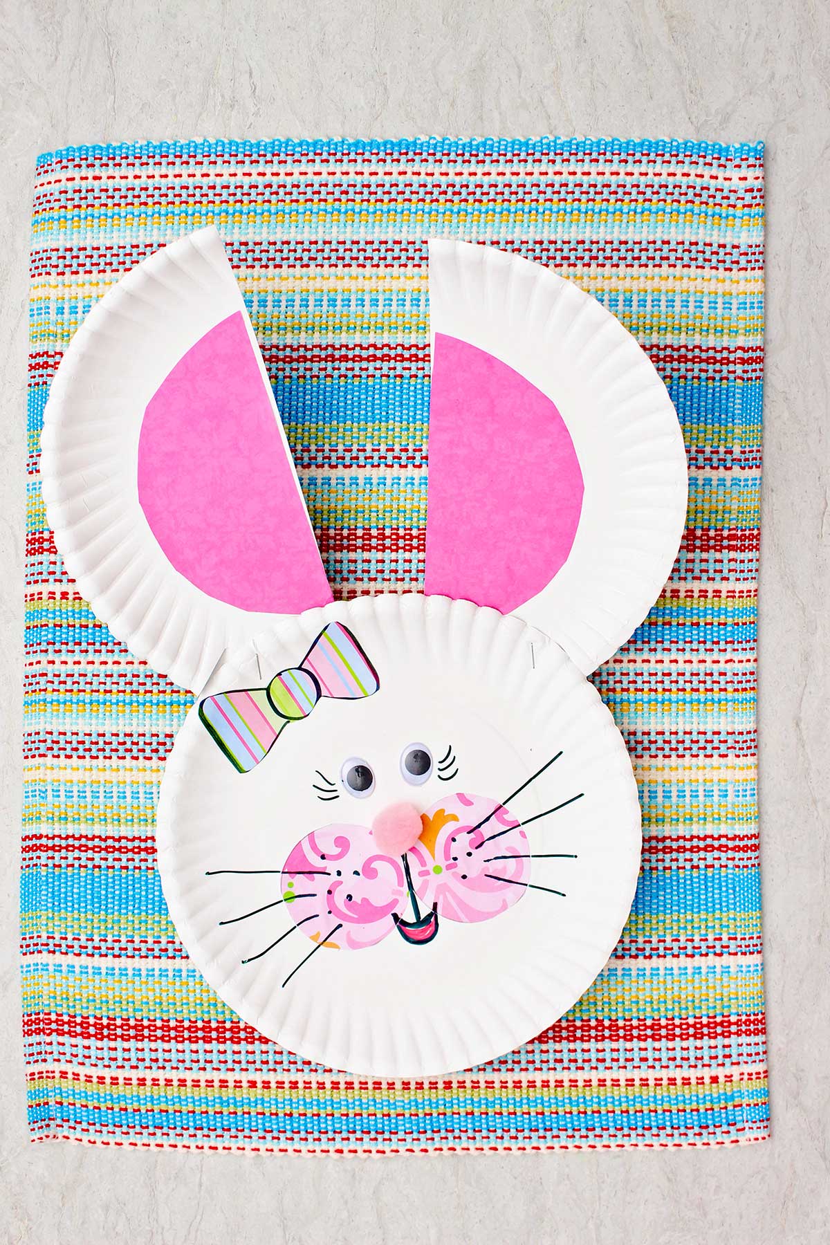 Coelhinho de prato de papel com orelhas e bochechas rosas, nariz de pom pom, laço listrado e olhos googly.