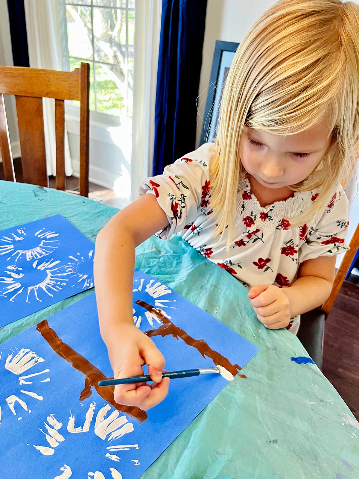 Menina loira com camisa floral adiciona tinta branca para neve em galhos em seu adorável artesanato de impressão de mão de cardeal de inverno.