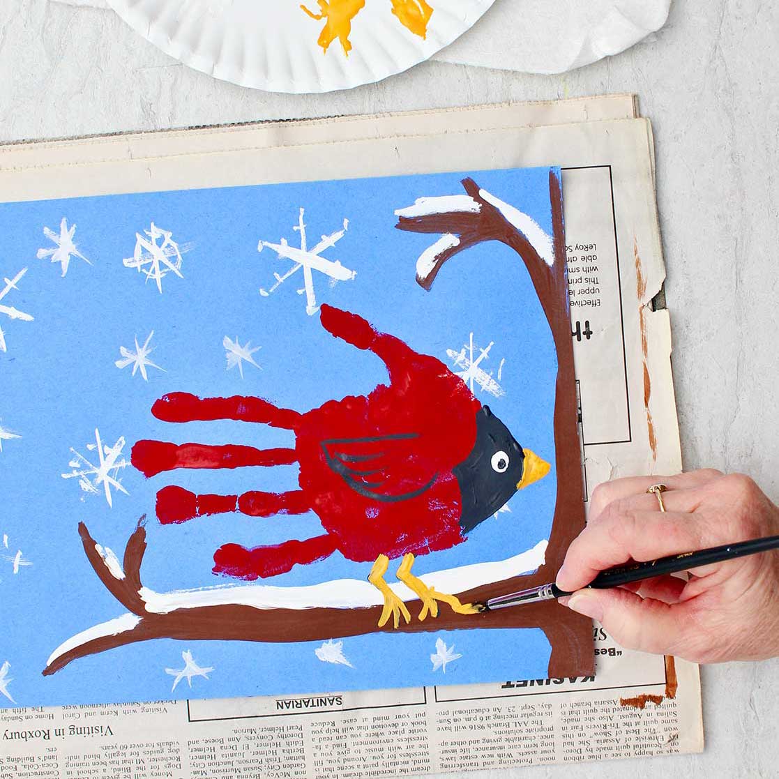 Pessoa pintando os detalhes finais em uma pintura de impressão de mão de cardeal inverno fofa de uma impressão de mão de cardeal sentada em um galho coberto de neve.