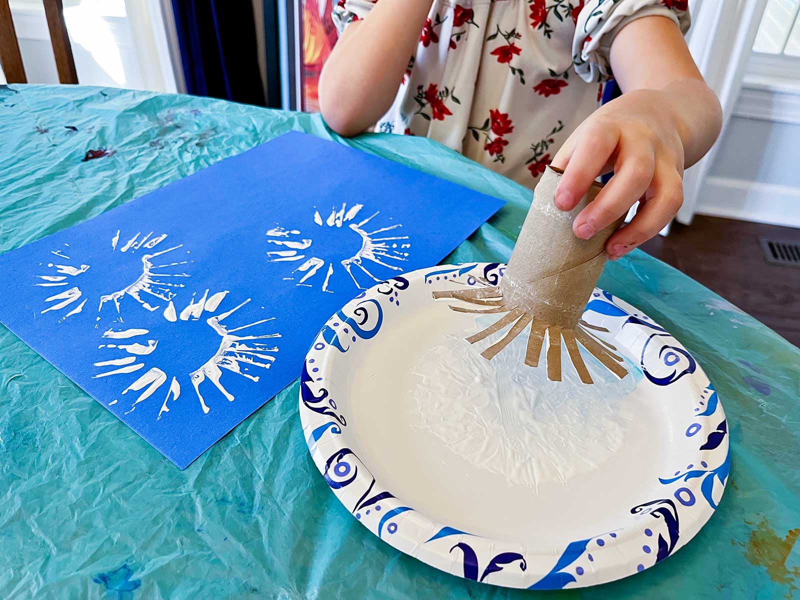 Menina pequena usando um rolo de toalha de papel cortado como um sol para fazer carimbos para seu projeto em uma folha de papel azul.