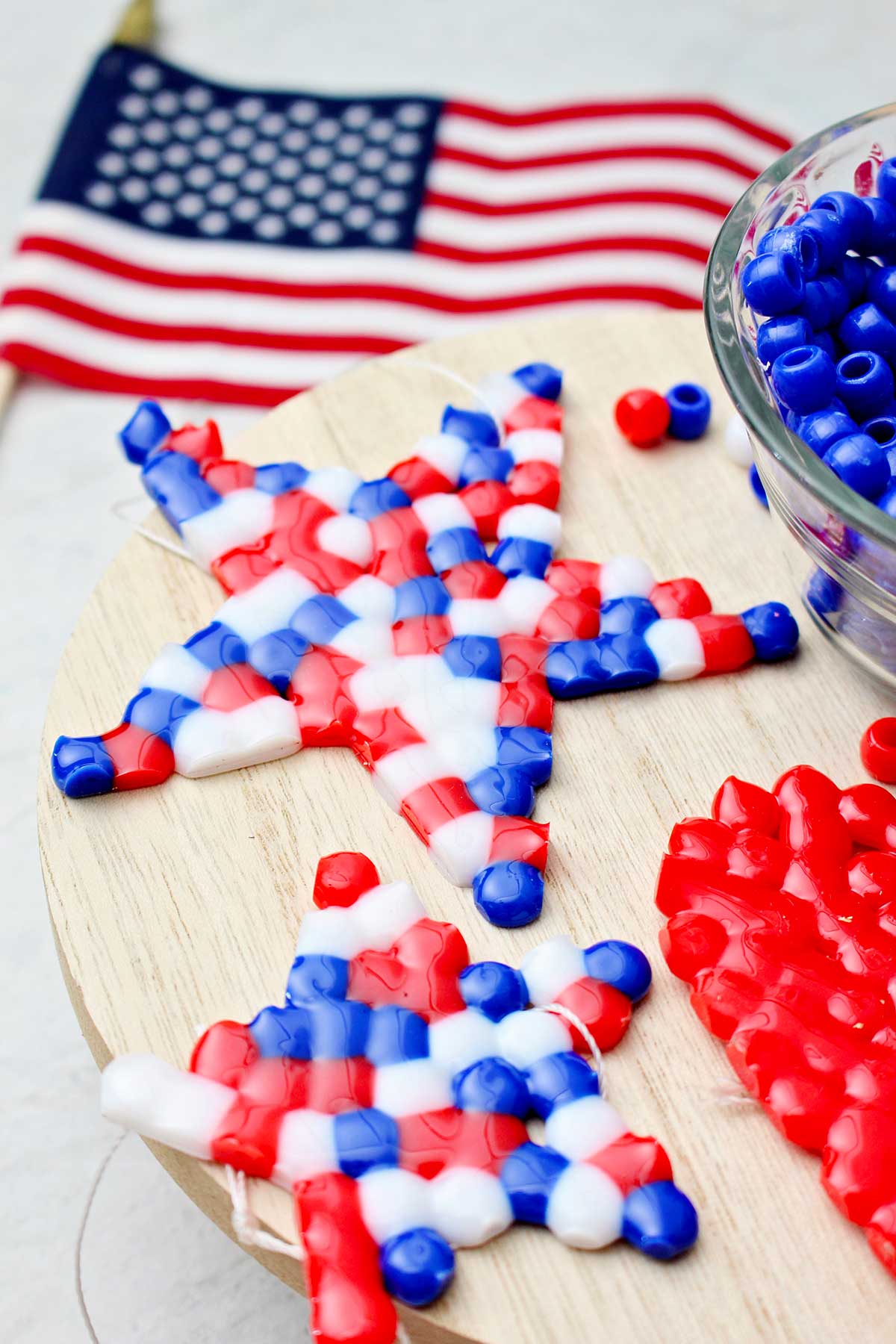 Duas pérolas derretidas concluídas, vermelha, branca e azul, em forma de estrela, sentadas sobre um pedaço circular de madeira ao lado de uma tigela de pérolas azuis com uma pequena bandeira americana ao fundo.