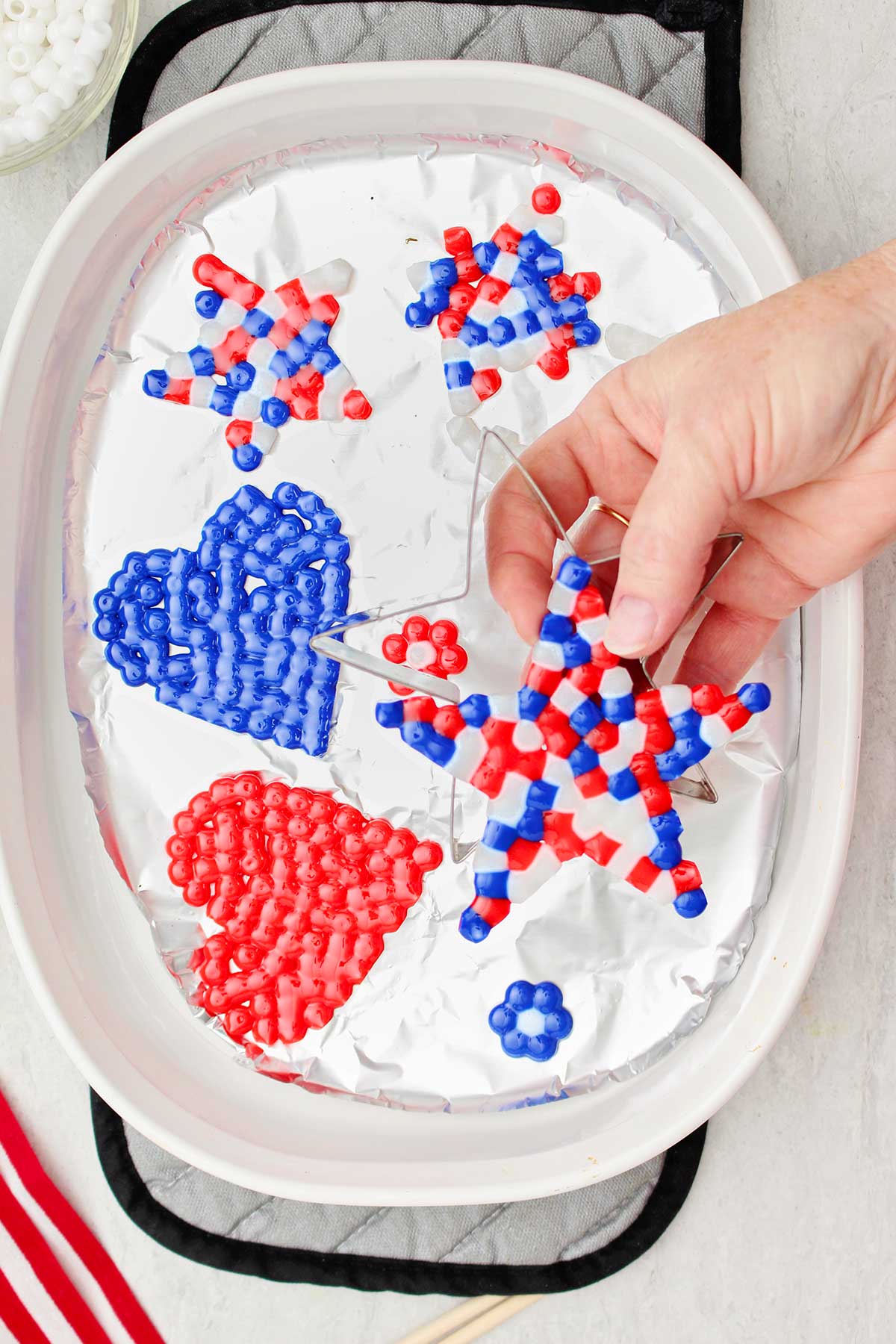 Uma pessoa retirando uma estrela de contas derretidas nas cores vermelha, branca e azul de um cortador de biscoitos em forma de estrela, com outros projetos de contas derretidas atrás.