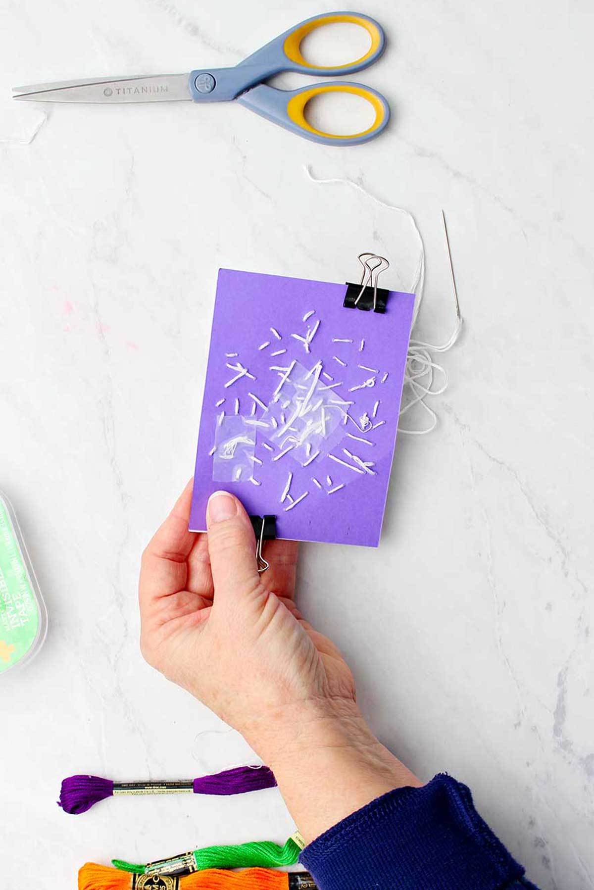 Mão segurando atrás do papel roxo mostrando os pontos de bordado.
