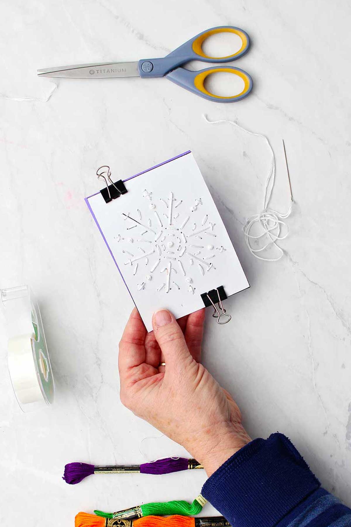 Mão segurando papel de floco de neve bordado preso em cartão roxo com fio de bordado, fita e tesoura próximas.
