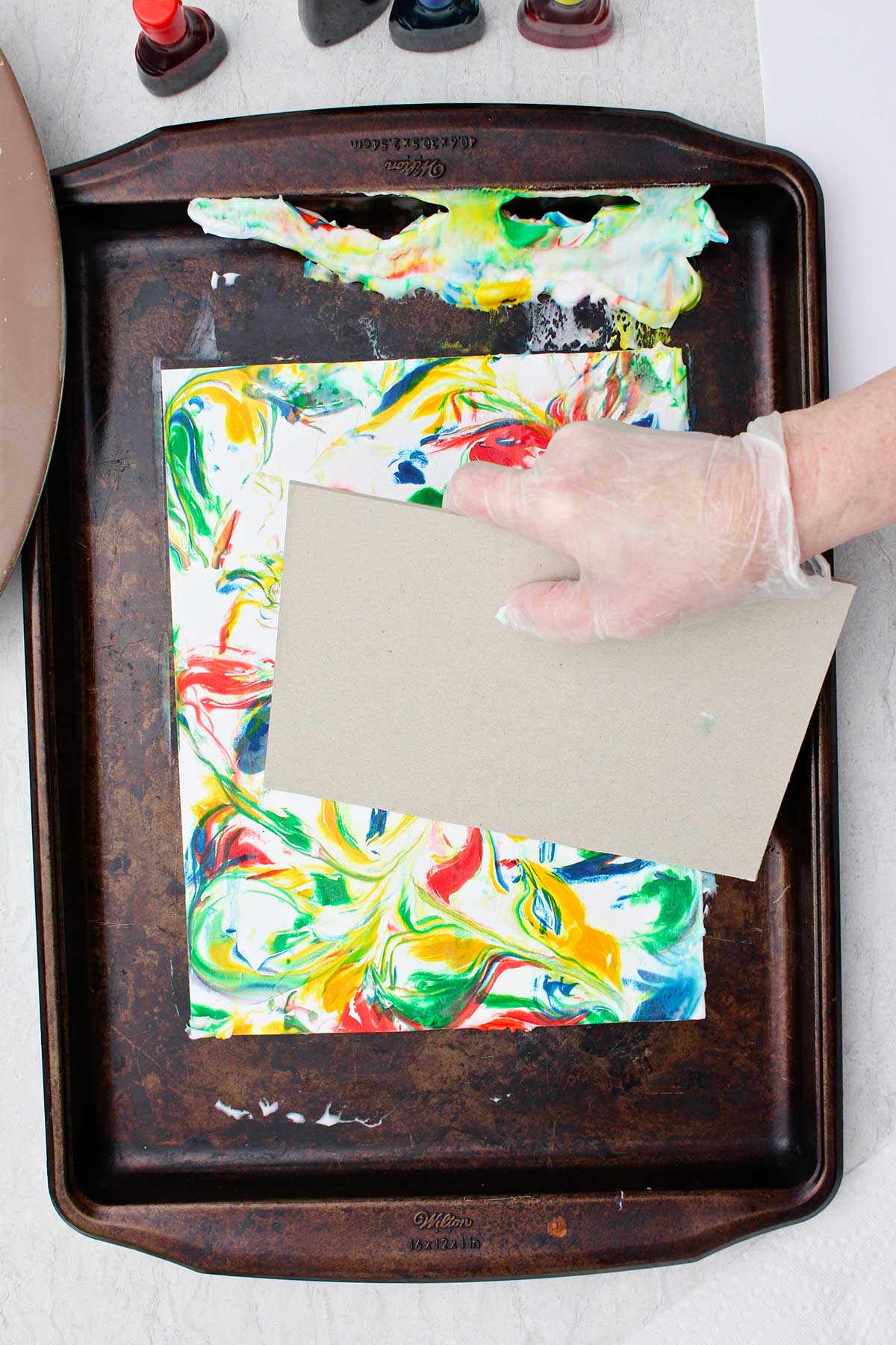 Pessoa usando um pedaço de papelão rígido para raspar o excesso de creme de barbear e corante alimentar em uma assadeira.