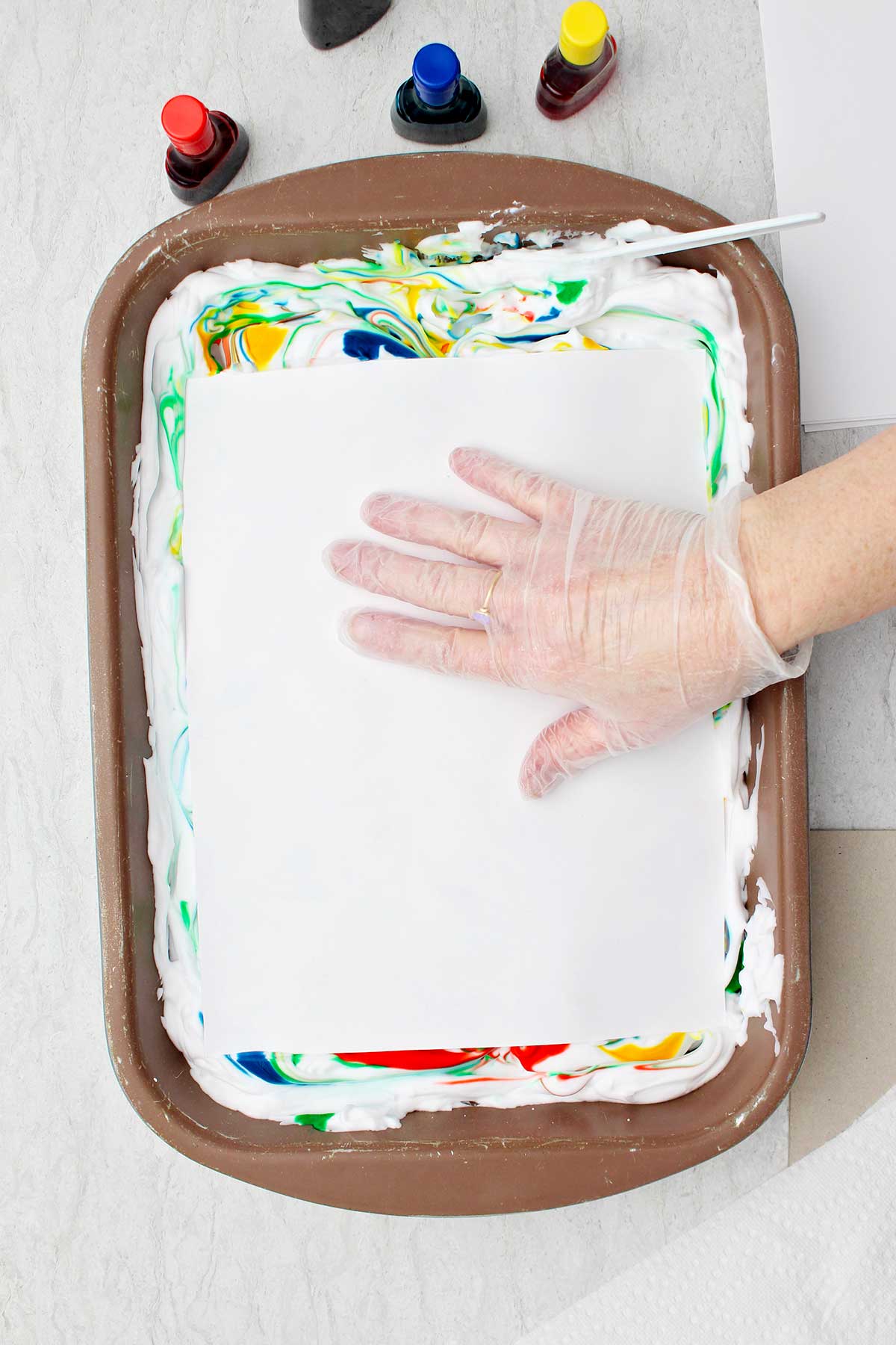 Pessoa usando luvas plásticas pressionando um pedaço de papel contra o creme de barbear colorido.