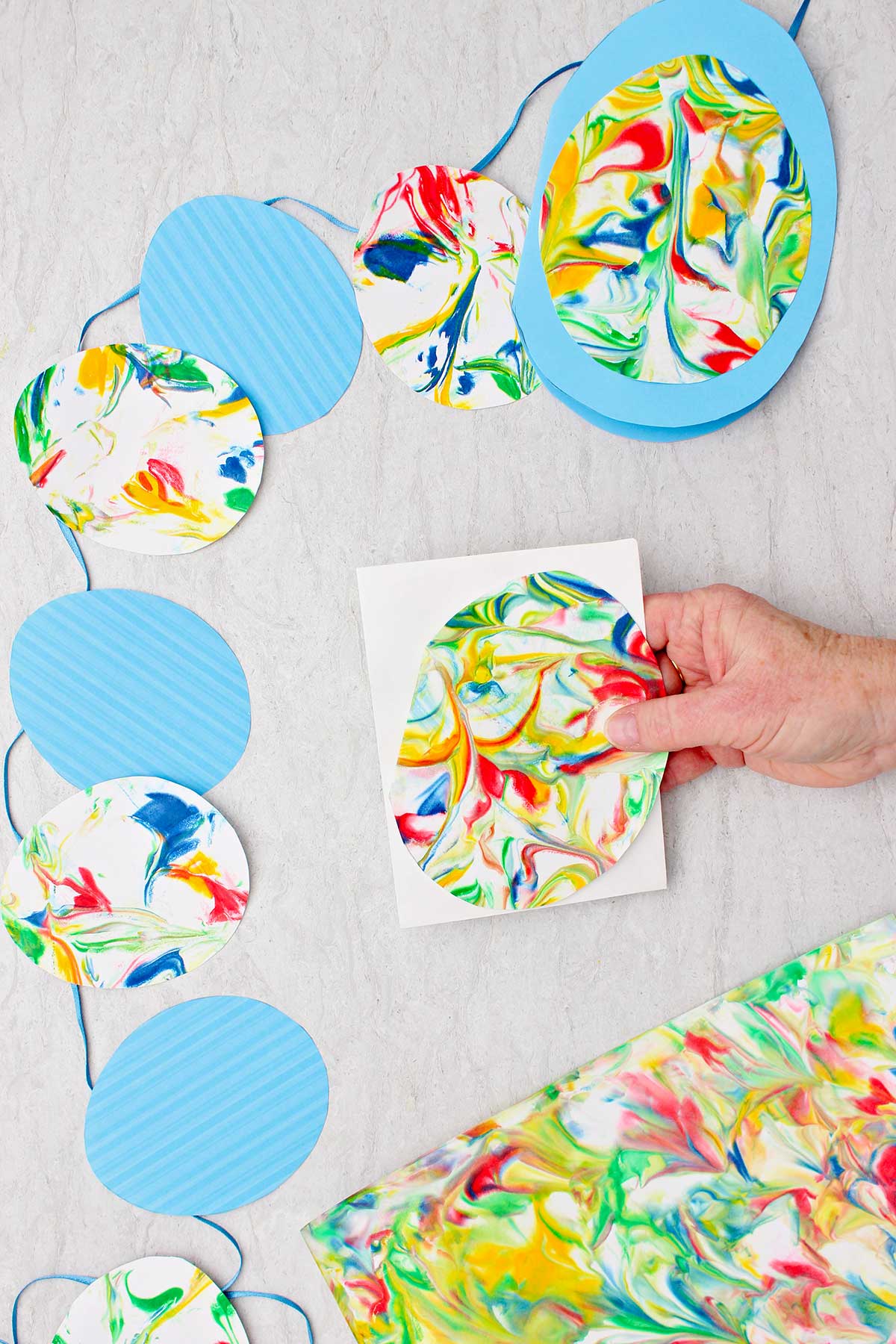 Pessoa segurando um ovo marmorizado sobre um pedaço de papel branco com bandeirinhas de ovos de Páscoa feitos de ovos marmorizados e ovos de papel azul por perto.