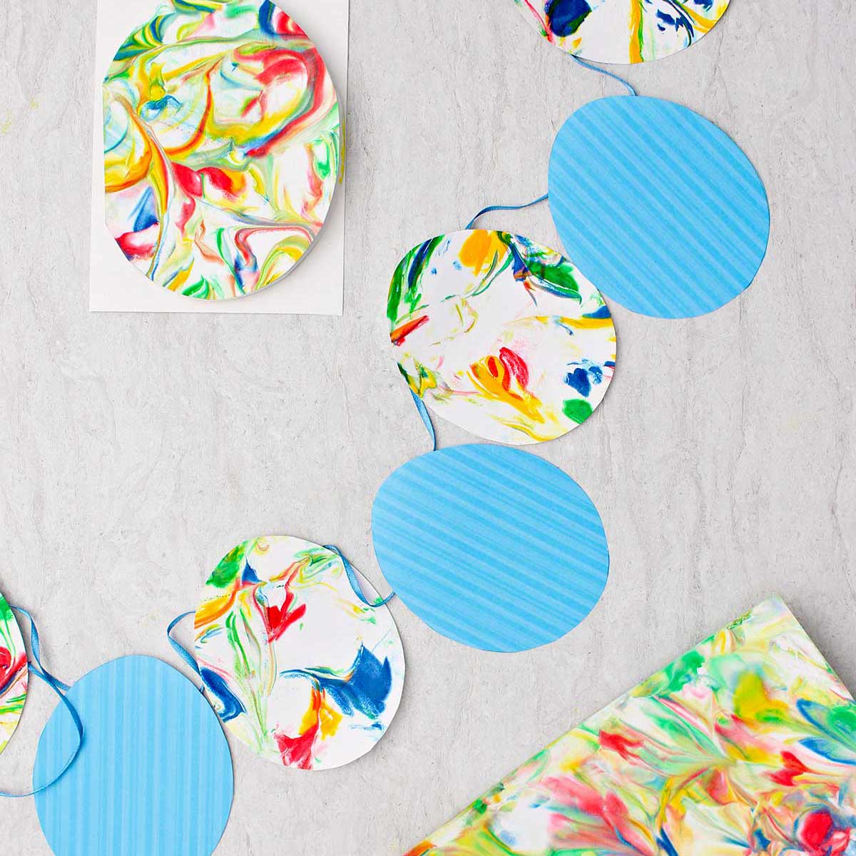 Bandeirinhas de ovo de Páscoa feitas com ovos de papel listrados azuis e ovos de papel marbrado em uma fita azul com outros pedaços de papel marbrado ao redor.