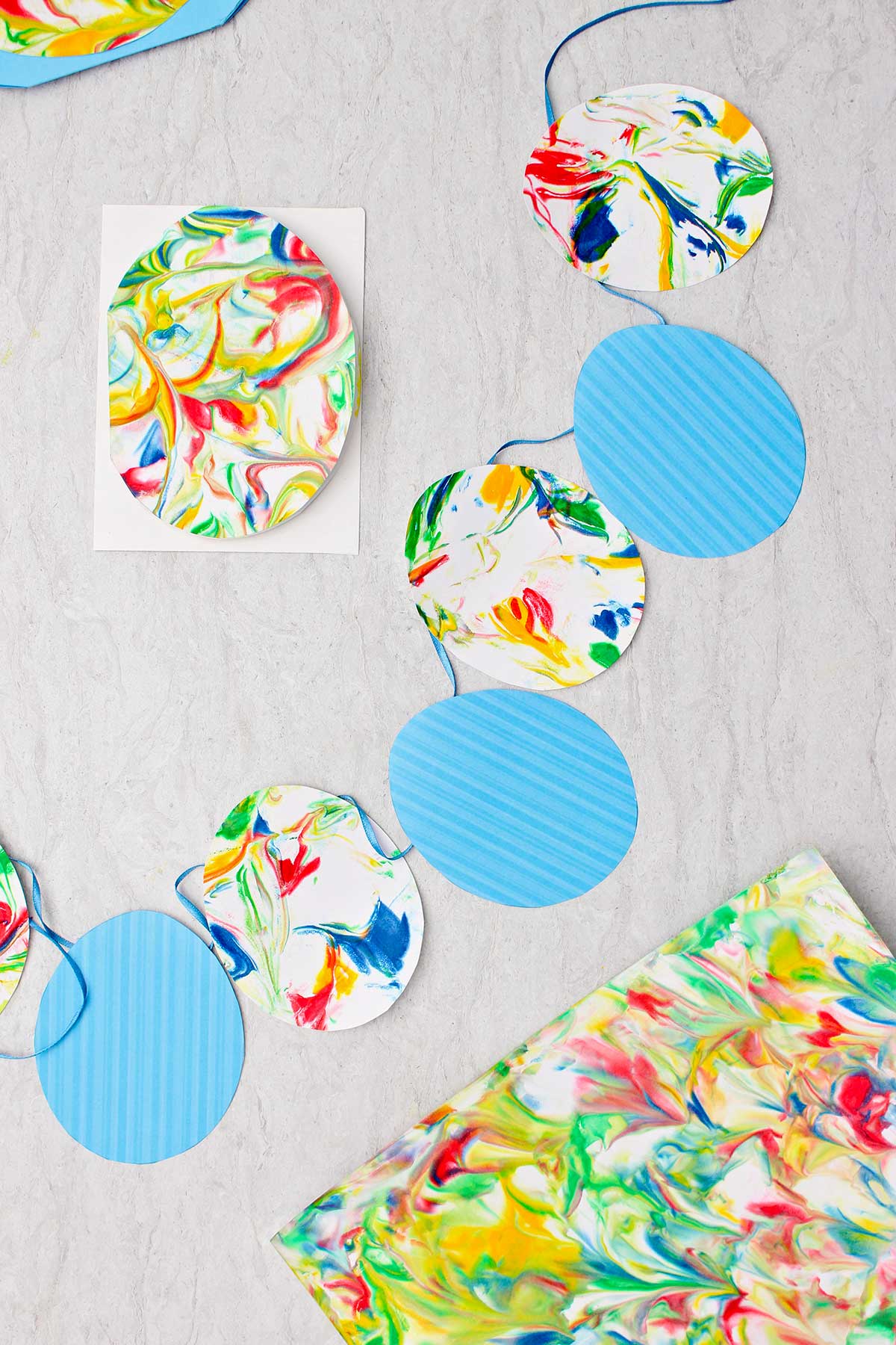 Bandeirinhas de ovo de Páscoa feitas com ovos de papel listrados azuis e ovos de papel marbrado em uma fita azul com outros pedaços de papel marbrado ao redor.