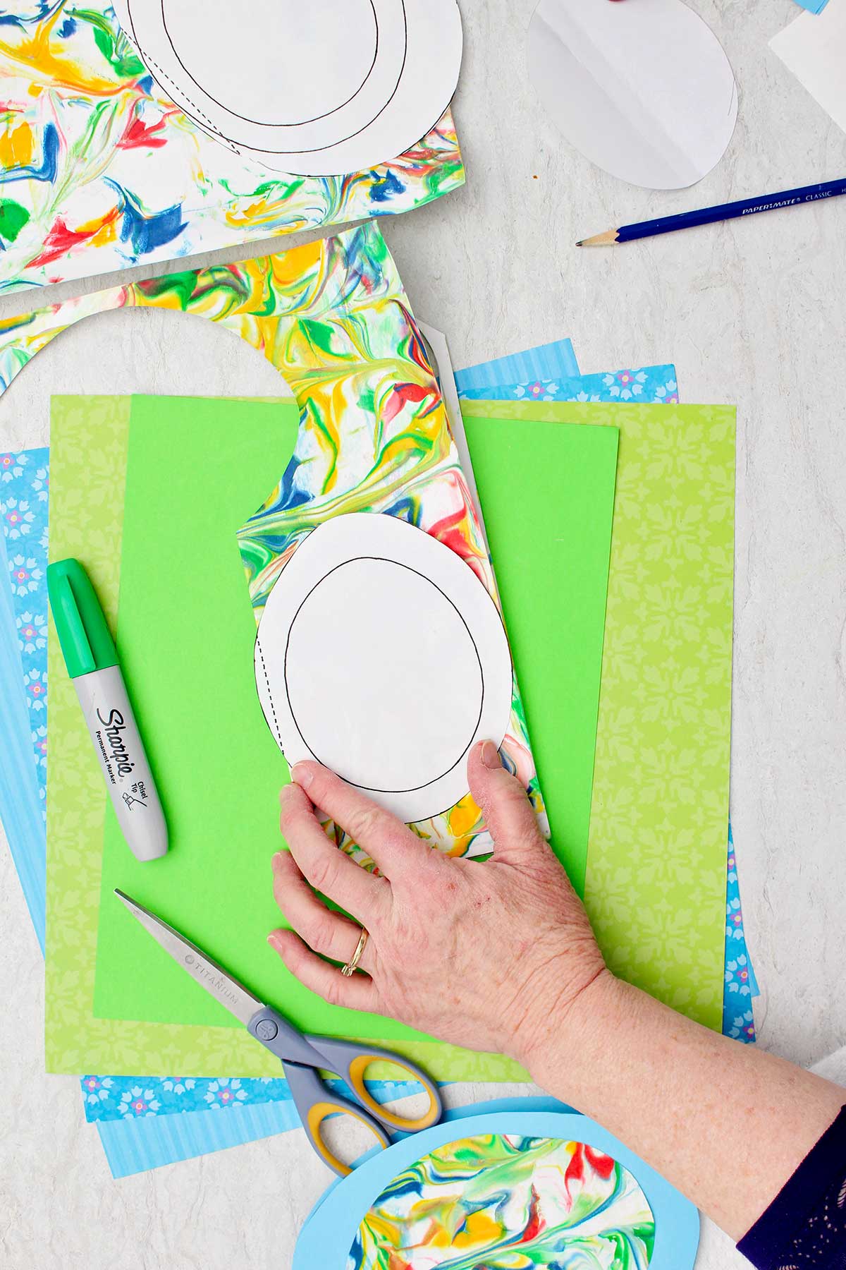 Pessoa traçando um molde de ovo em papel colorido mármore com outros materiais próximos.