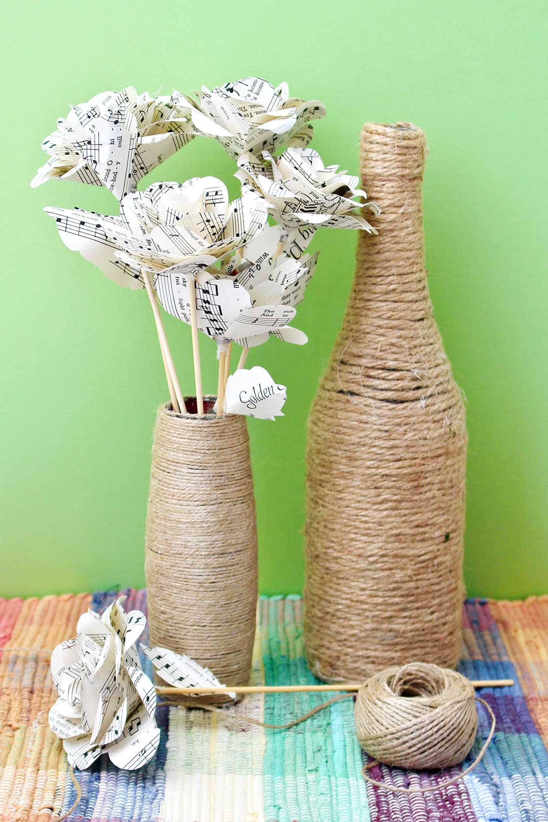 Uma garrafa envolta em barbante e um vaso envolto em barbante com flores de papel dentro, em uma toalha de mesa colorida contra um fundo verde.