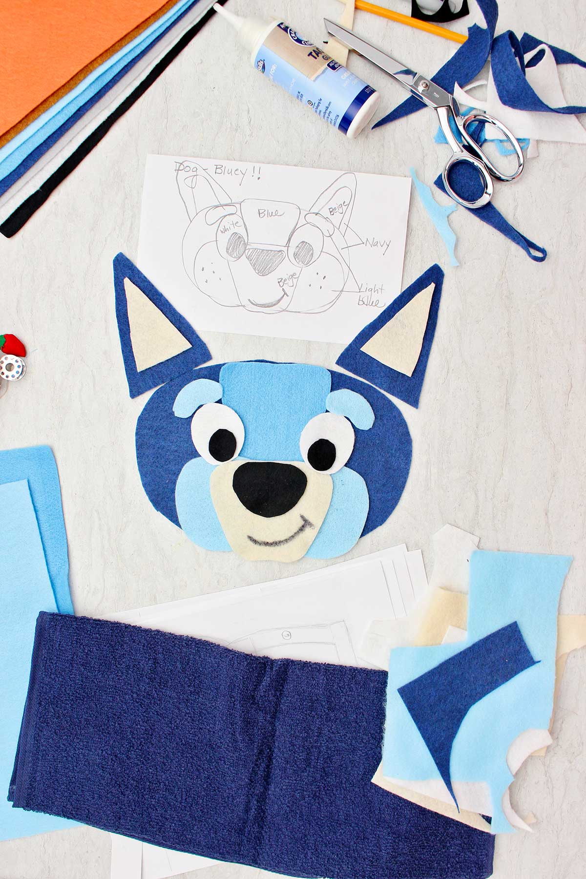 Um esboço para uma fantasia do Bluey próximo a alguns pedaços cortados de feltro para o rosto e outros retalhos de materiais ao redor.