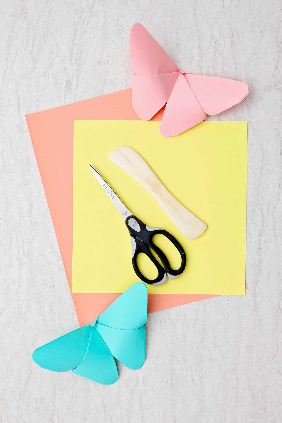 Uma borboleta de origami rosa e azul claro, um pedaço de papel amarelo e laranja, um vincador de papel de madeira e uma tesoura.