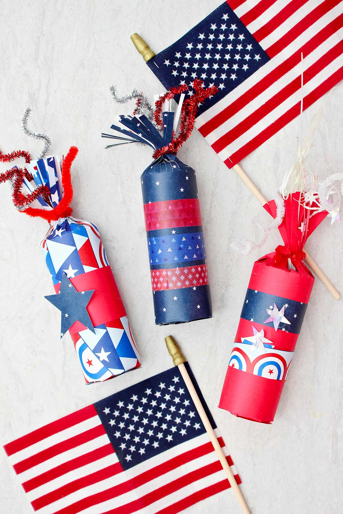 Três caixas de guloseimas em forma de fogos de artifício decoradas com papéis vermelhos, brancos e azuis e fita washi, com duas pequenas bandeiras americanas como decoração.