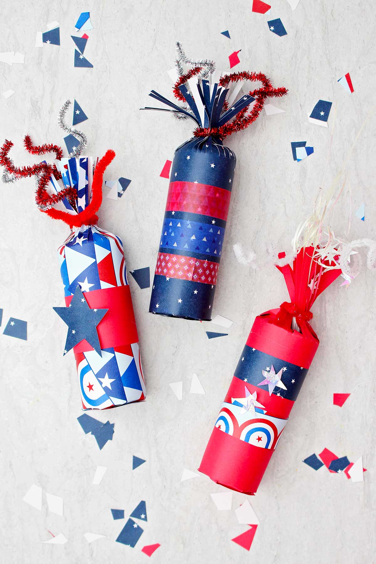 Três caixas de guloseimas de foguete decoradas com papel vermelho, branco e azul e fita washi, com alguns pedaços de papel festivo espalhados ao redor.