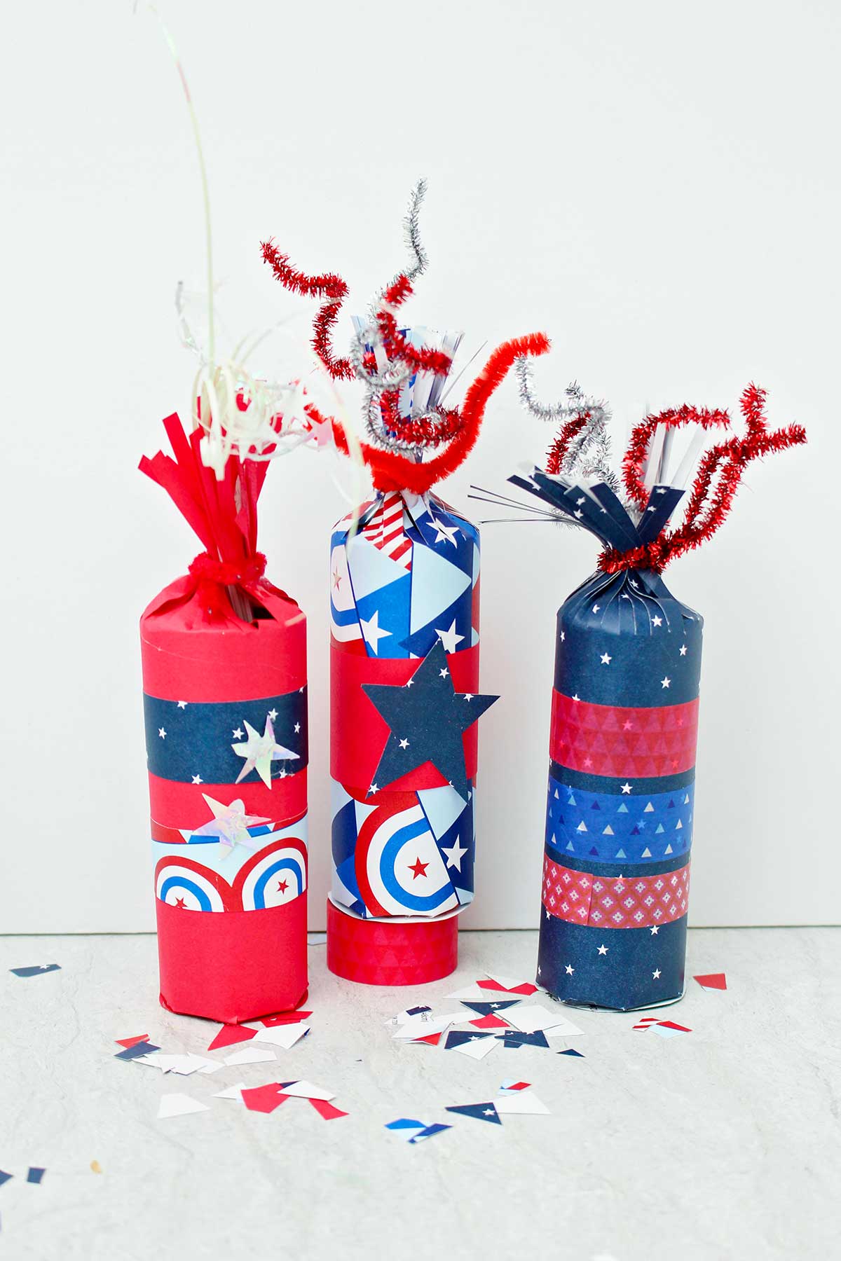 Vista direta de três caixas de guloseimas em forma de fogos de artifício decoradas com papéis vermelhos, brancos e azuis e fita washi, com alguns retalhos de papel festivo espalhados ao redor.