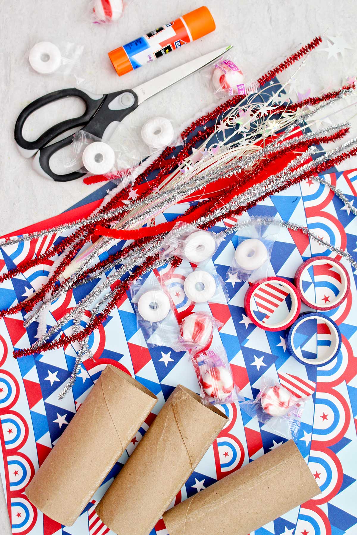 Fitas de pipe cleaner brilhantes, papel de scrapbooking vermelho, branco e azul, fita washi vermelha e azul, rolos de papel toalha, tesoura, cola e balas para colocar nas caixas de guloseimas.