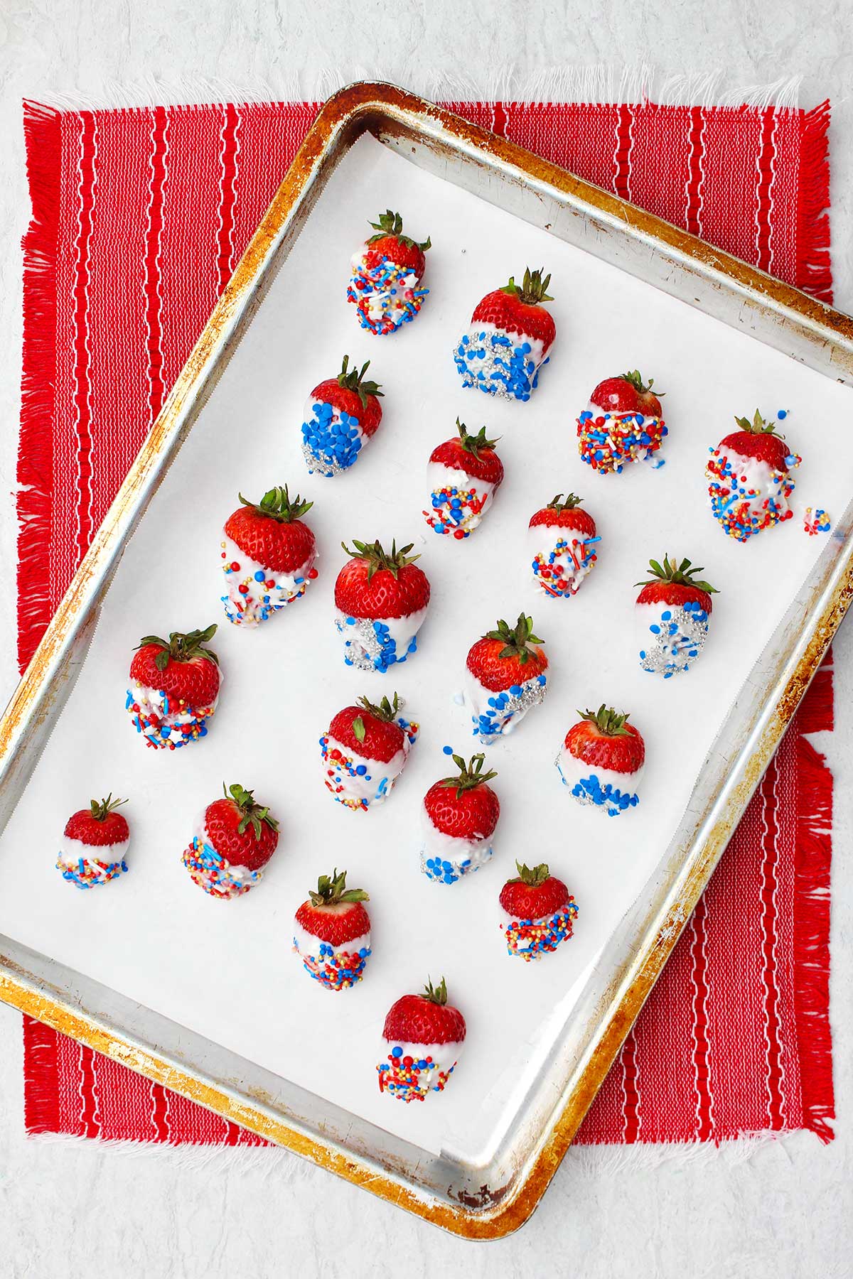 Morangos mergulhados em chocolate branco completos com confetes azuis, vermelhos e prateados em uma assadeira com papel manteiga.